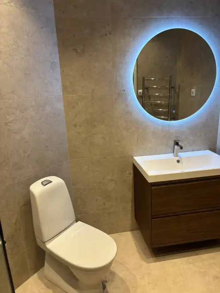 Renoverat badrum på Valhallavägen med WC, träfärgad kommod och rund belyst spegel.