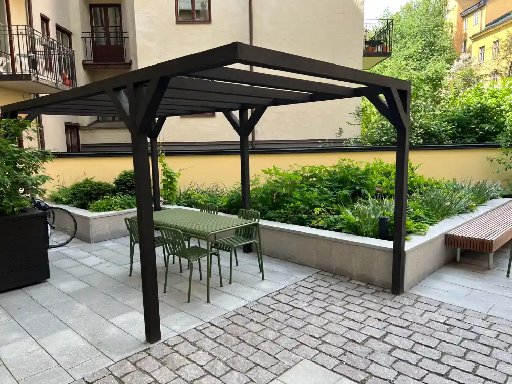 Svart pergola med bord och stolar på en stenlagd innergård hos BRF Kastanjen på Södermalm efter innergårdsrenovering.