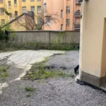 Innergård med äldre markplattor, grus och växtlighet vid entrén före renovering hos BRF Kastanjen på Södermalm.